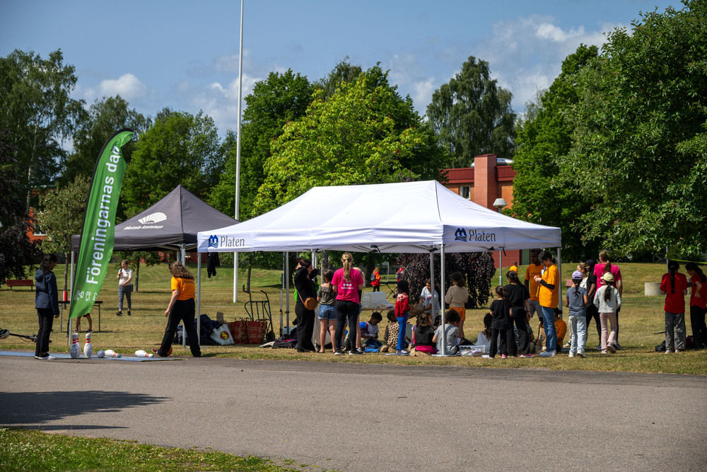 Många barn och vuxna ledare under ett vitt tält i ett bostadsområde, med grönska kring.