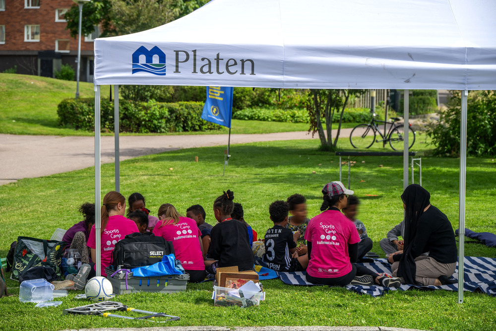 Många barn och ledare i färgglada rosa tröjor under ett vitt tält på en gräsmatta